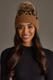 A woman with long brown hair smiles against a plain background, wearing the Panache Apparel Collection’s Camel Leopard Pattern Knit Pom Hat, a brown beanie featuring black leopard print and a faux fur pom-pom, paired with a ribbed black top.