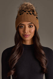 A woman with long brown hair is wearing the Panache Apparel Collection’s Camel Leopard Pattern Knit Pom Hat and a black ribbed long-sleeve top, standing against a plain gray background.