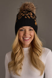 A woman with long blonde hair smiles against a plain gray background, wearing a white ribbed turtleneck and the Panache Apparel Collection Black Leopard Pattern Knit Pom Hat, featuring a leopard print design and faux fur pom-pom.