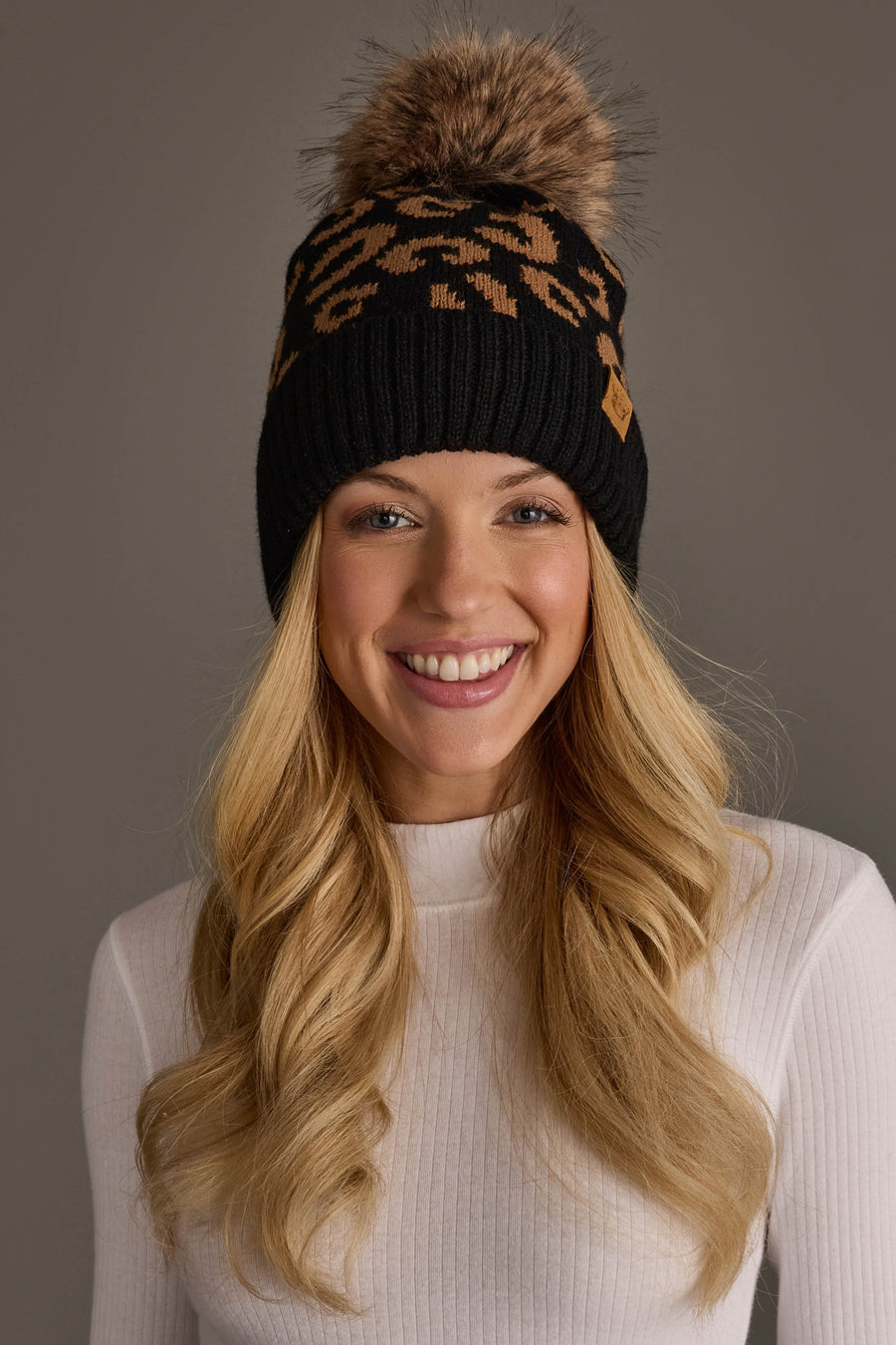 A woman with long blonde hair smiles against a plain gray background, wearing a white ribbed turtleneck and the Panache Apparel Collection Black Leopard Pattern Knit Pom Hat, featuring a leopard print design and faux fur pom-pom.