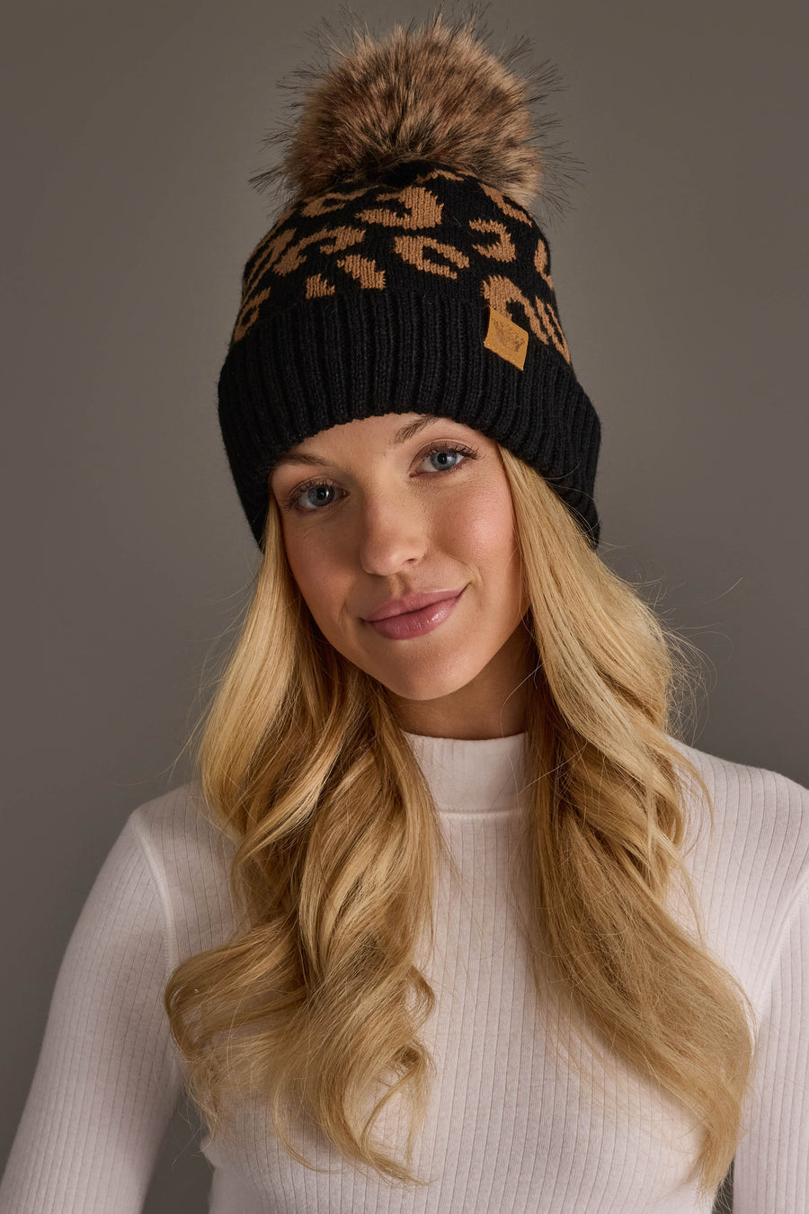 A woman with long blonde hair wears the Panache Apparel Collection Black Leopard Pattern Knit Pom Hat, featuring a faux fur pom-pom, and a white ribbed top, standing against a plain grey background.