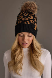 A woman with long blonde hair wears a white ribbed turtleneck and the Black Leopard Pattern Knit Pom Hat by Panache Apparel Collection, a black knit beanie featuring a brown leopard print design and a large faux fur pom-pom, while standing against a plain gray background.