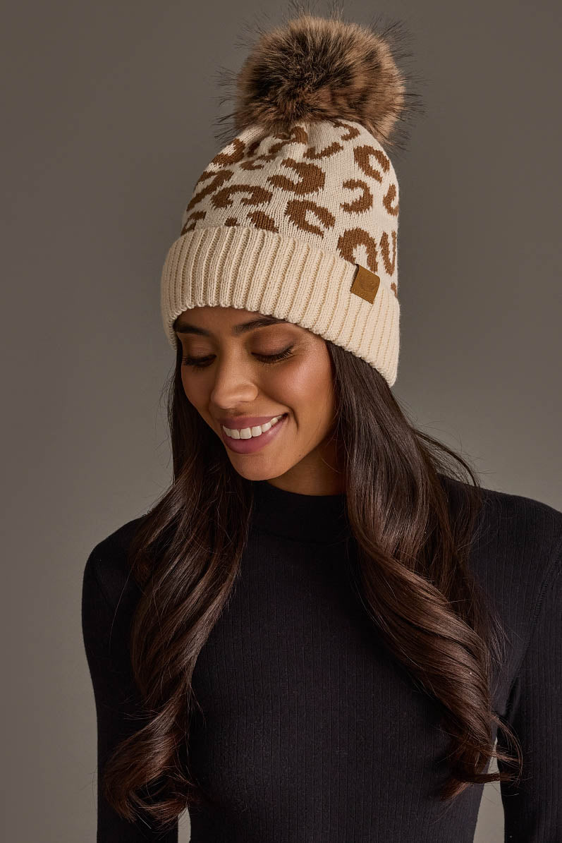A woman with long brown hair smiles while looking down, wearing the Panache Apparel Collection Cream Leopard Pattern Knit Pom Hat, a cream knit beanie featuring a brown leopard print and a faux fur pom-pom, paired with a black ribbed top against a plain gray background.