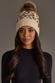A woman with long brown hair is wearing the Cream Leopard Pattern Knit Pom Hat from Panache Apparel Collection, a cream knit beanie featuring a brown leopard print and a furry pom-pom, paired with a black ribbed top, standing against a plain gray background.