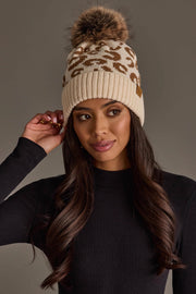 A woman wearing the Cream Leopard Pattern Knit Pom Hat by Panache Apparel Collection, a beige knit beanie with a brown leopard print and a fluffy pom pom, pairs it with a long-sleeve black ribbed shirt, gazing to the side while adjusting her hat.