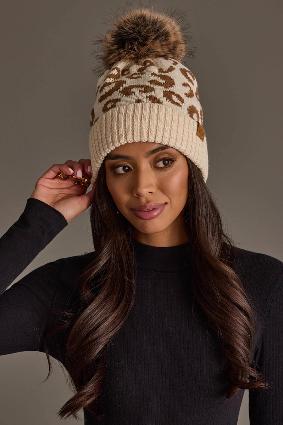 A woman wearing the Cream Leopard Pattern Knit Pom Hat by Panache Apparel Collection, a beige knit beanie with a brown leopard print and a fluffy pom pom, pairs it with a long-sleeve black ribbed shirt, gazing to the side while adjusting her hat.