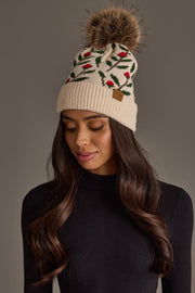 A woman wearing the Cream Floral Pattern Knit Pom Hat from Panache Apparel Collection, featuring red and green leaf designs and a large faux fur pom-pom, pairs it with a black ribbed turtleneck as she looks downward against a neutral background.