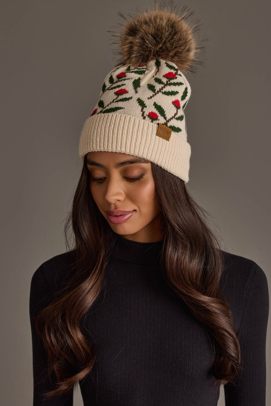 A woman wearing the Cream Floral Pattern Knit Pom Hat from Panache Apparel Collection, featuring red and green leaf designs and a large faux fur pom-pom, pairs it with a black ribbed turtleneck as she looks downward against a neutral background.