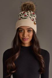 A woman with long, wavy dark hair models the Panache Apparel Collection’s Cream Floral Pattern Knit Pom Hat, featuring red and green embroidery and a brown faux fur pom-pom, paired with a black ribbed turtleneck top against a neutral background.