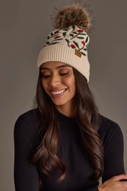 A woman with long brown hair is wearing the Cream Floral Pattern Knit Pom Hat by Panache Apparel Collection, featuring a faux fur pompom. She smiles and looks down, dressed in a black top.