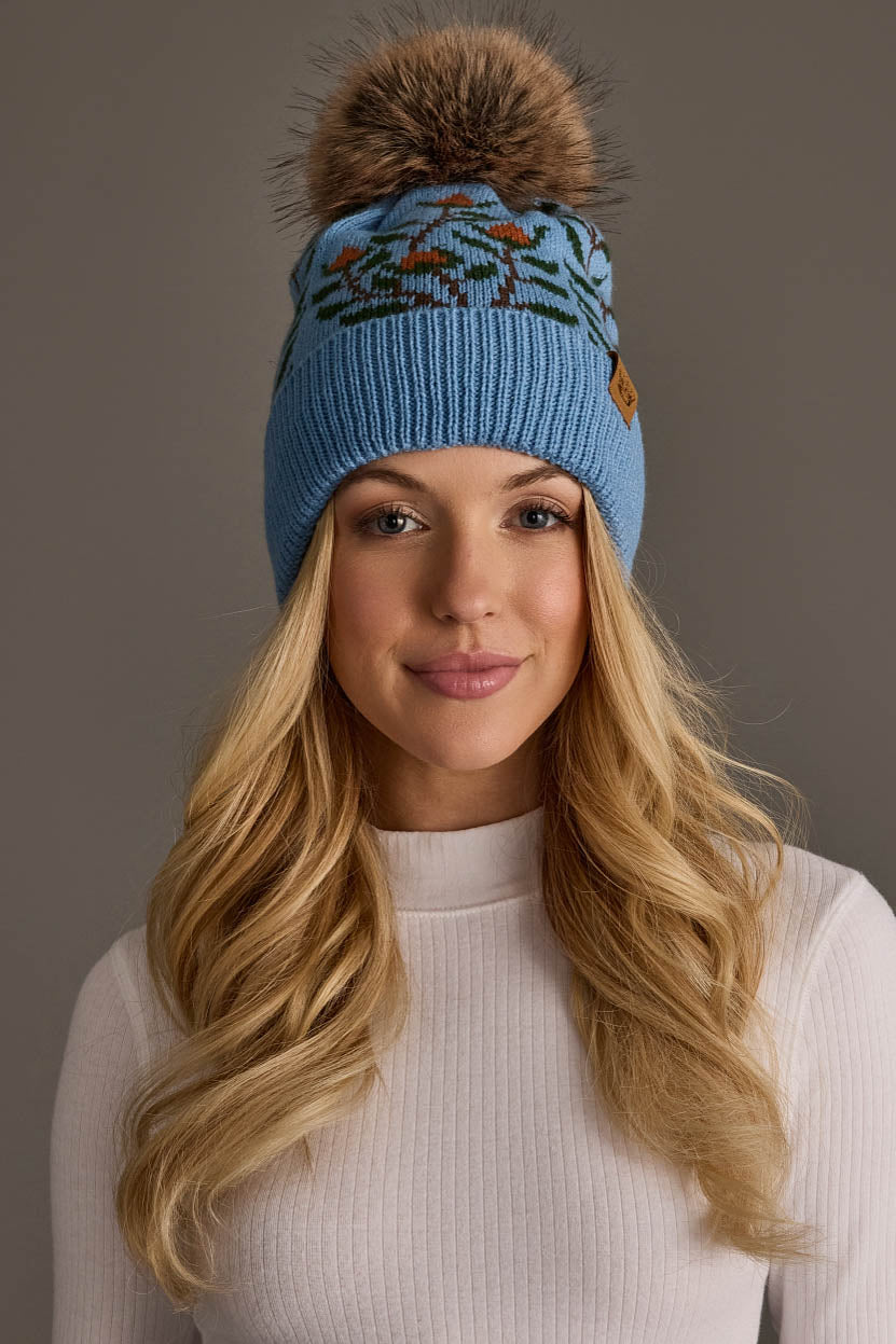 A woman with long blonde hair is wearing the Panache Apparel Collection’s Blue Floral Pattern Knit Pom Hat, featuring a fluffy pom-pom and intricate design. She’s dressed in a fitted white turtleneck and smiling at the camera against a neutral background.