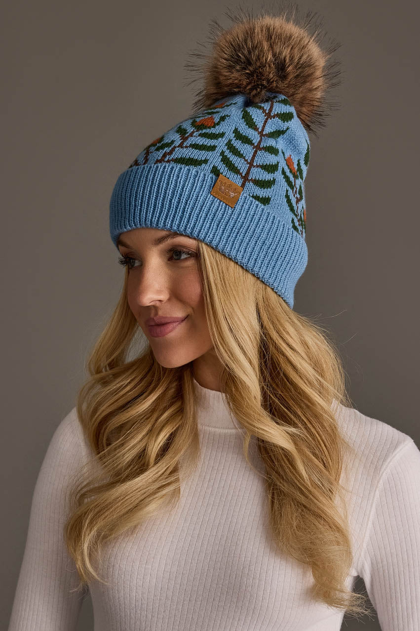 A woman with long blonde hair is wearing the Panache Apparel Collection’s Blue Floral Pattern Knit Pom Hat and a white ribbed sweater, looking to the side against a plain background.