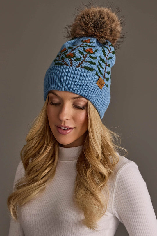 A woman with long blonde hair is wearing the Panache Apparel Collection Blue Floral Pattern Knit Pom Hat and a white ribbed turtleneck top, standing against a plain gray background.