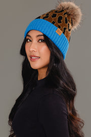 A woman with long dark hair models the Panache Apparel Collection Brown Leopard Pattern Pom Hat with Blue Cuff, featuring a faux fur pom-pom, styled with a black top against a plain gray background.