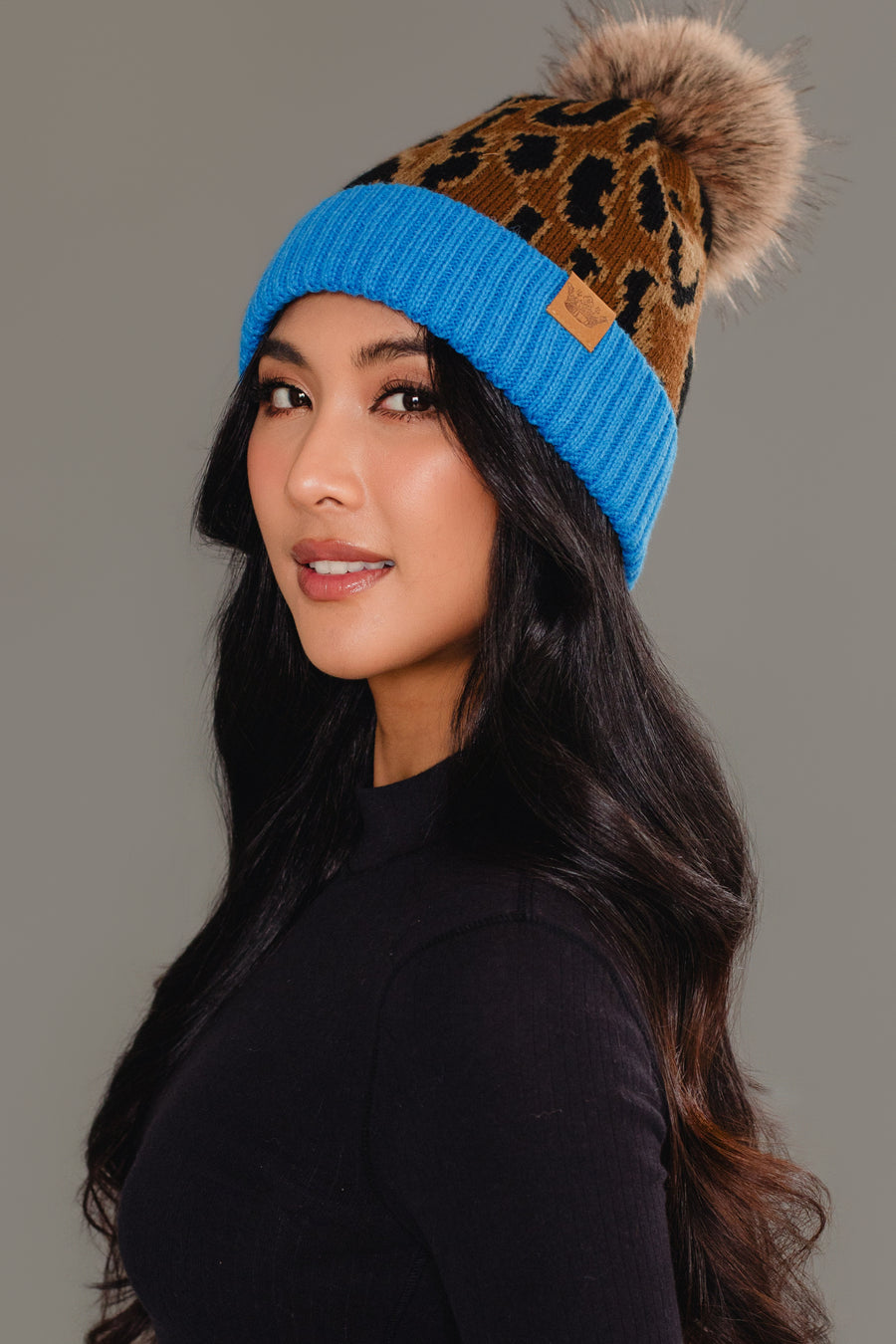 A woman with long dark hair models the Panache Apparel Collection Brown Leopard Pattern Pom Hat with Blue Cuff, featuring a faux fur pom-pom, styled with a black top against a plain gray background.