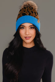 A woman with long black hair wearing a black top and the Panache Apparel Collection Brown Leopard Pattern Pom Hat with Blue Cuff, featuring a brown leopard print design, blue brim, and brown pom-pom, against a plain gray background.