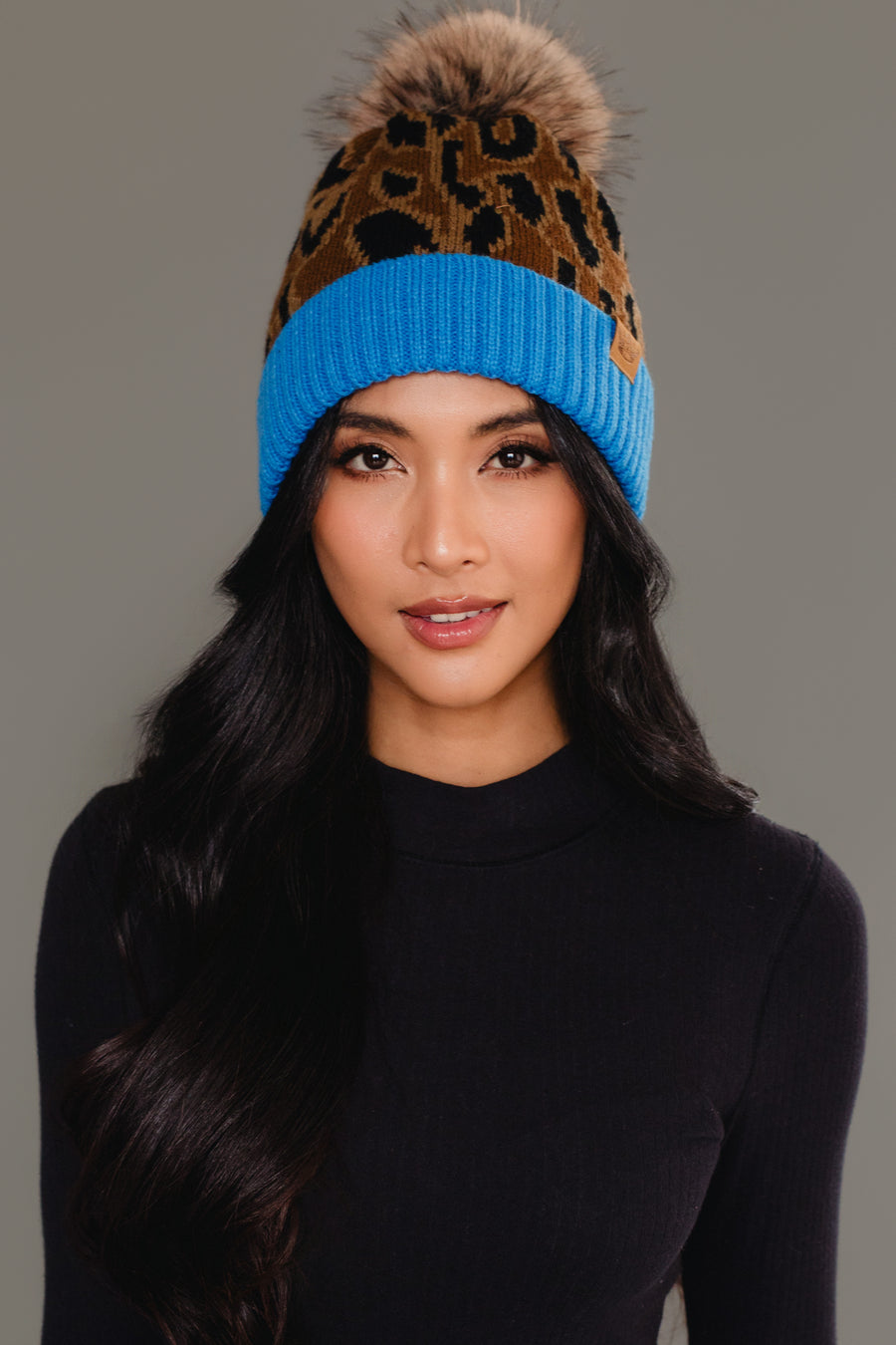 A woman with long black hair wearing a black top and the Panache Apparel Collection Brown Leopard Pattern Pom Hat with Blue Cuff, featuring a brown leopard print design, blue brim, and brown pom-pom, against a plain gray background.