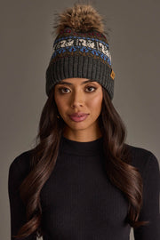 A woman with long, wavy dark hair wears a black knit sweater and the Panache Apparel Collection’s Gray Swan Pattern Knit Pom Hat, featuring a patterned band and a brown faux fur pom-pom, as she stands against a plain, neutral background.