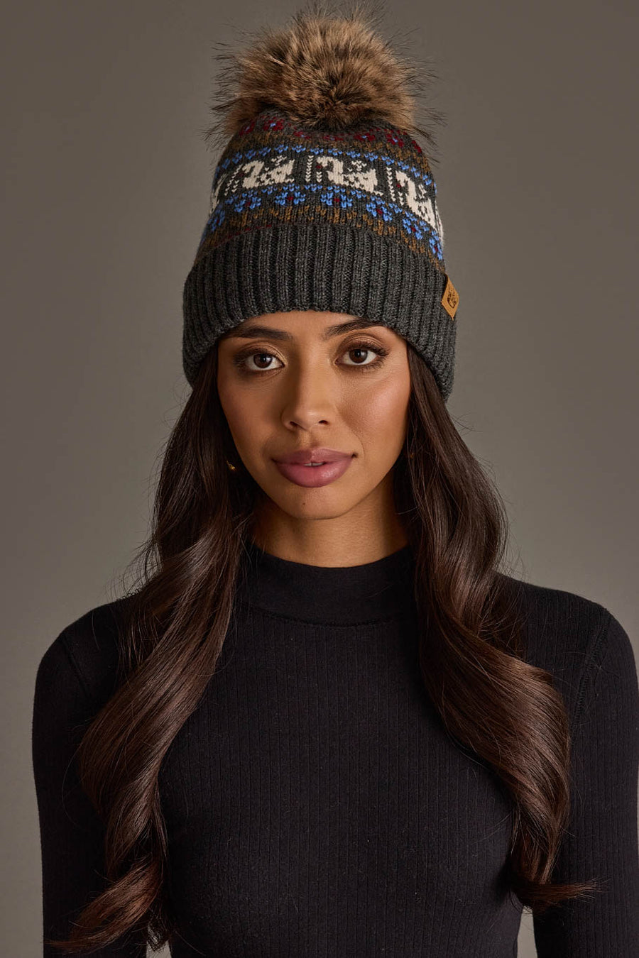 A woman with long, wavy dark hair wears a black knit sweater and the Panache Apparel Collection’s Gray Swan Pattern Knit Pom Hat, featuring a patterned band and a brown faux fur pom-pom, as she stands against a plain, neutral background.