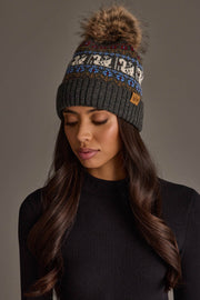 A woman with long dark hair is wearing the Panache Apparel Collection’s Gray Swan Pattern Knit Pom Hat and a black long-sleeve top, standing against a plain background.