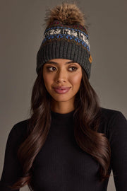 A woman with long wavy hair, wearing a black knit sweater and the Gray Swan Pattern Knit Pom Hat from Panache Apparel Collection, a gray winter hat featuring a blue-and-white pattern and topped with a pom-pom, looks slightly to the side.