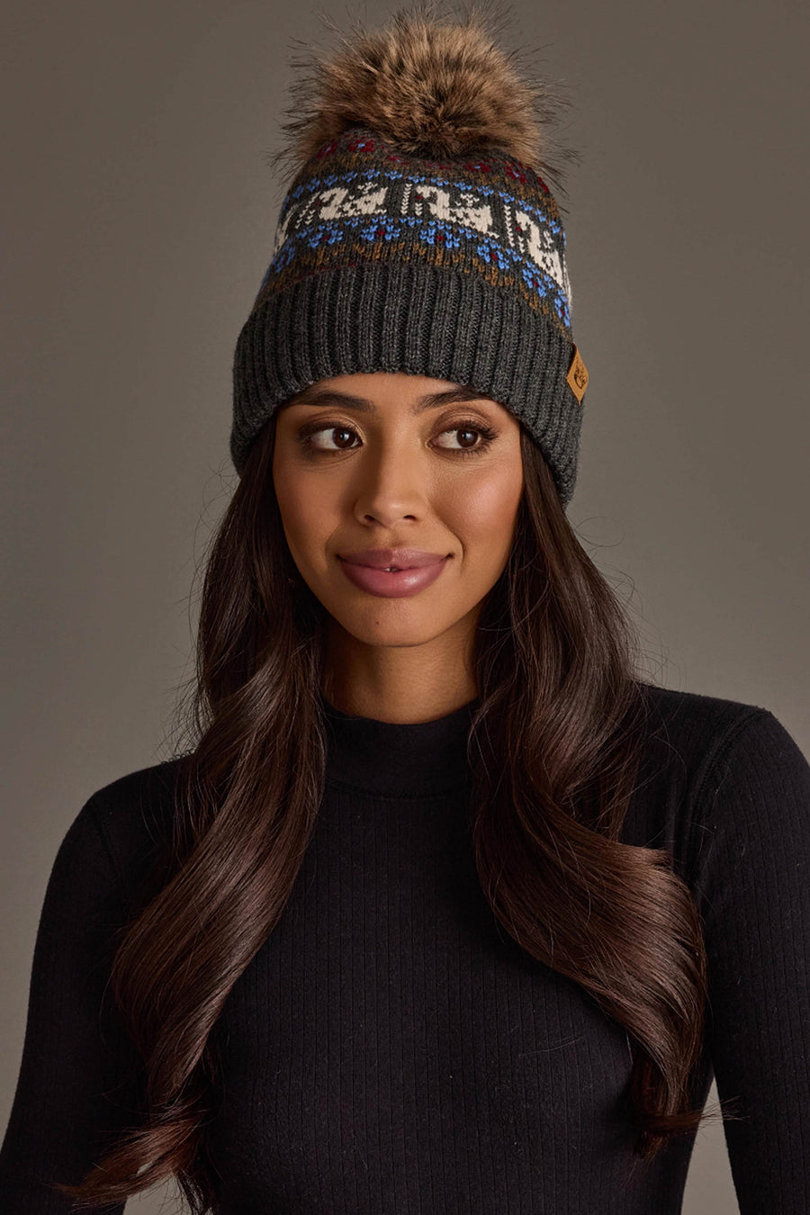 A woman with long wavy hair, wearing a black knit sweater and the Gray Swan Pattern Knit Pom Hat from Panache Apparel Collection, a gray winter hat featuring a blue-and-white pattern and topped with a pom-pom, looks slightly to the side.