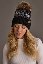 A woman with long blonde hair is wearing the Panache Apparel Collection Black Swan Pattern Knit Pom Hat featuring colorful patterns and a brown faux fur pom-pom, along with a white ribbed top, looking down with a neutral expression against a gray background.