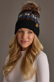 A woman with long blonde hair wears the Panache Apparel Collection Black Swan Pattern Knit Pom Hat featuring colorful patterns and a fur pom-pom, paired with a white ribbed long-sleeve top, as she looks slightly to the side and smiles softly.