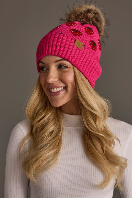 A smiling woman with long blonde hair wears the Panache Apparel Collection “Strawberry Pattern Pom Hat,” a bright pink knit beanie featuring red sequin lips and a brown faux fur pom-pom, paired with a white ribbed turtleneck sweater.