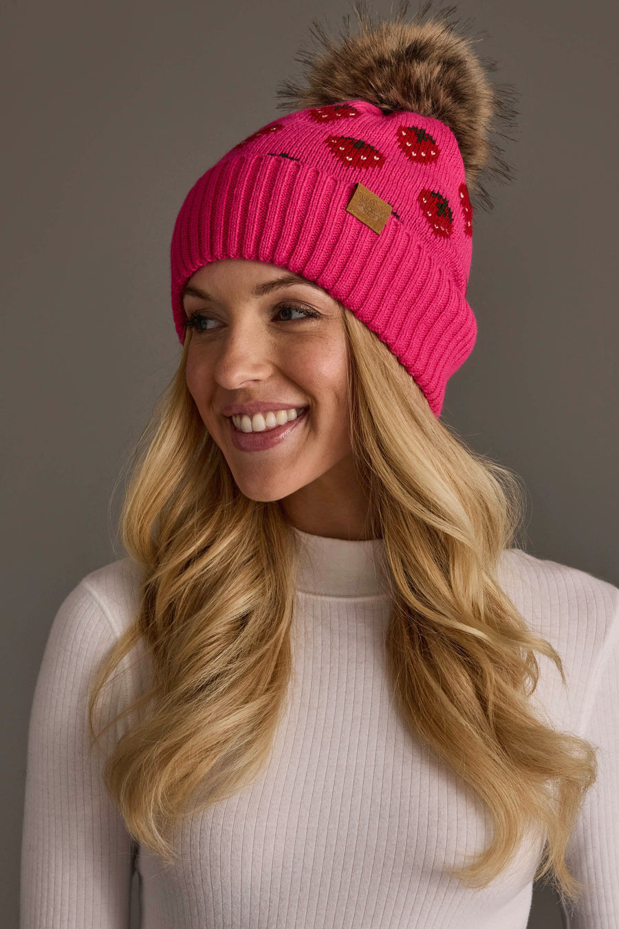 A smiling woman with long blonde hair wears the Panache Apparel Collection “Strawberry Pattern Pom Hat,” a bright pink knit beanie featuring red sequin lips and a brown faux fur pom-pom, paired with a white ribbed turtleneck sweater.