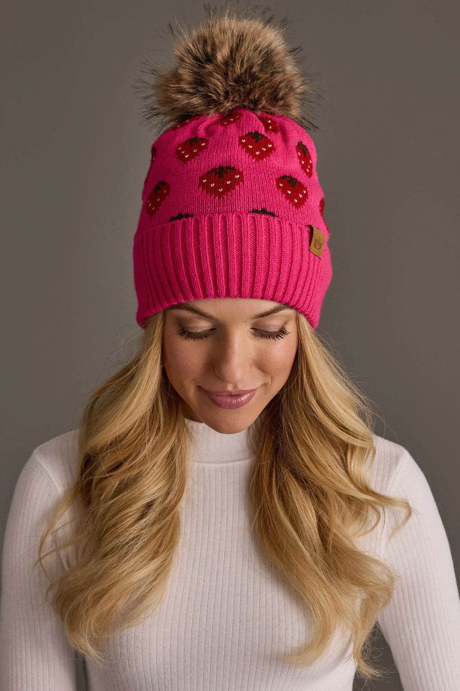A woman with long blonde hair wears the Panache Apparel Collection’s Strawberry Pattern Pom Hat, a bright pink knit hat featuring red lips and a brown faux fur pom-pom, paired with a white ribbed turtleneck, as she looks down against a plain gray background.