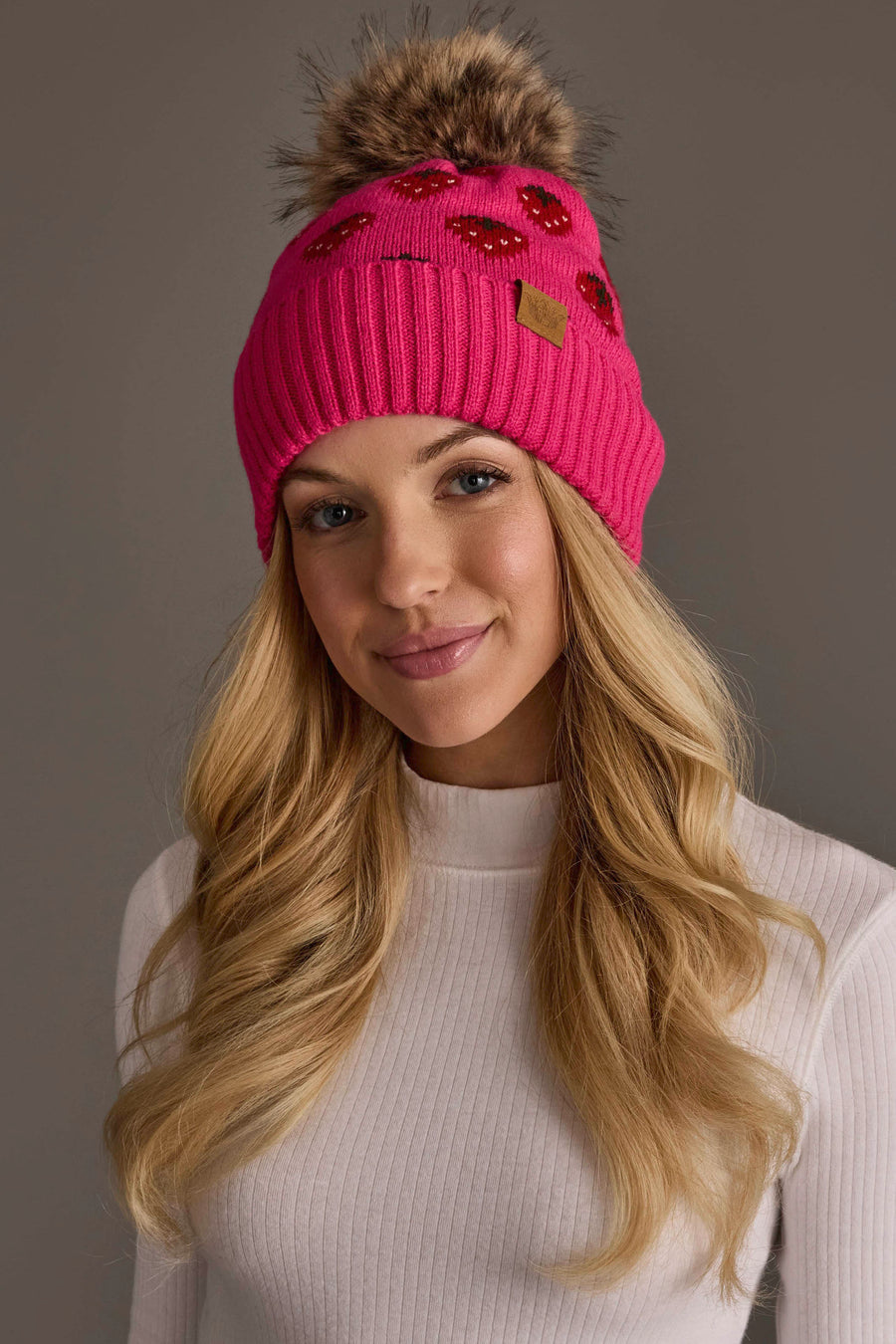 A woman with long blonde hair wears the Panache Apparel Collection’s Strawberry Pattern Pom Hat, a bright pink knit beanie featuring a faux fur pom-pom and sequin detail, paired with a white ribbed turtleneck sweater as she stands against a neutral gray background.