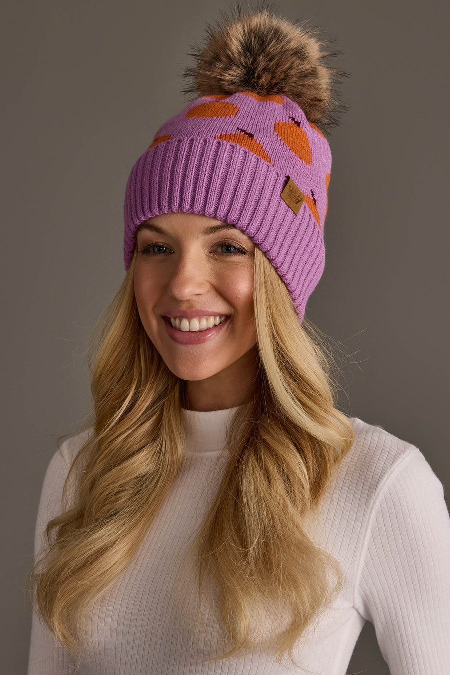 A smiling woman with long blonde hair wears the Panache Apparel Collection’s Orange Pattern Pom Hat, a purple knit beanie featuring orange shapes and a large fur pom-pom, paired with a ribbed white sweater, posing against a plain gray background.