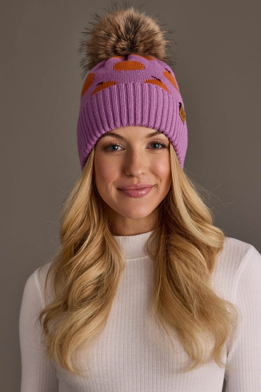 A woman with long blonde hair smiles while wearing the Panache Apparel Collection’s Orange Pattern Pom Hat, a purple knit beanie adorned with orange spots and topped with a large faux fur pom-pom, paired with a white ribbed top against a plain background.