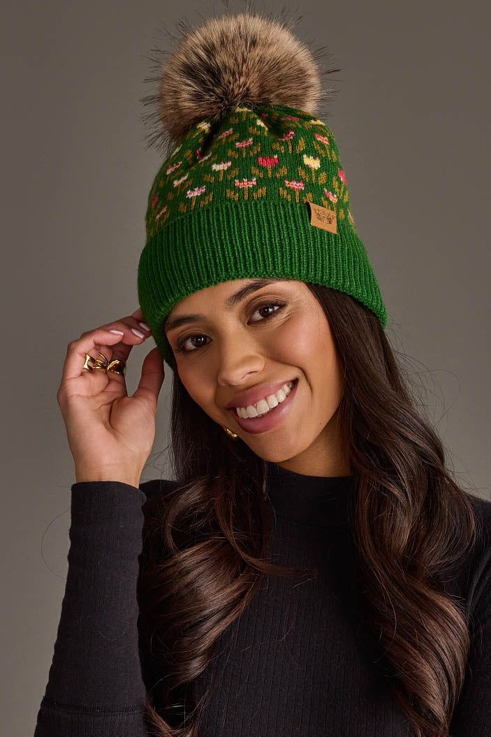 A woman with long, wavy brown hair smiles while touching the side of her Panache Apparel Collection Green Small Floral Pattern Knit Pom Hat, which features colorful patterns and a large faux fur pom-pom. She is also wearing a black long-sleeve top.