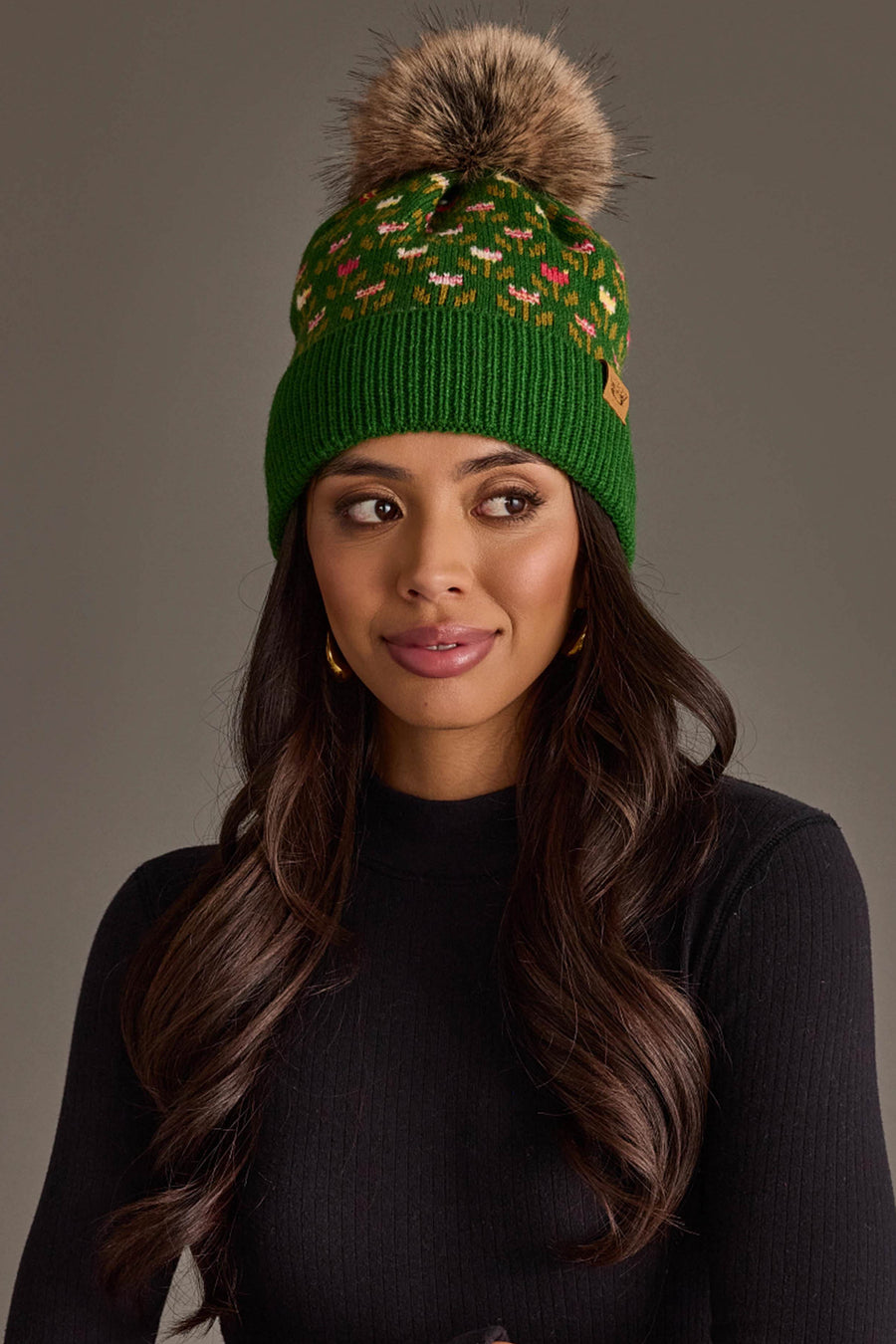 A woman with long brown hair is wearing the Panache Apparel Collection’s Green Small Floral Pattern Knit Pom Hat, paired with a black sweater and gold hoop earrings, looking slightly to the side against a plain background.