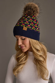 A woman with long blonde hair wears a white ribbed top and the Panache Apparel Collection Navy Small Floral Pattern Knit Pom Hat, which features colorful patterns and a large brown faux fur pom-pom, as she looks down with a slight smile.