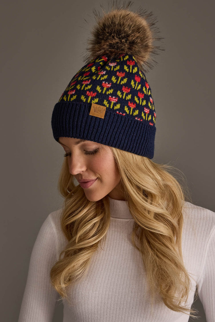 A woman with long blonde hair wears a white ribbed top and the Panache Apparel Collection Navy Small Floral Pattern Knit Pom Hat, which features colorful patterns and a large brown faux fur pom-pom, as she looks down with a slight smile.