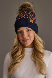 A young woman with long blonde hair is wearing a white ribbed sweater and the Navy Small Floral Pattern Knit Pom Hat by Panache Apparel Collection, featuring colorful patterns and a faux fur pom-pom, while standing against a plain gray background.