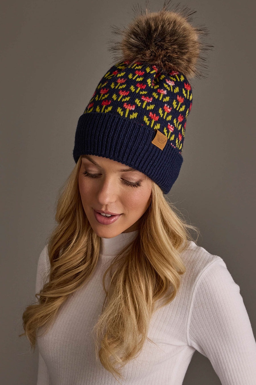 A woman with long blonde hair, wearing a white ribbed sweater and the Panache Apparel Collection Navy Small Floral Pattern Knit Pom Hat featuring a large brown faux fur pom-pom on top, looks slightly downward with a soft expression.