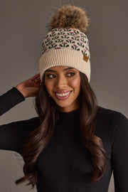A woman in a black sweater wears the Panache Apparel Collection Cream Small Floral Pattern Knit Pom Hat, smiling and touching her hair against a neutral background.