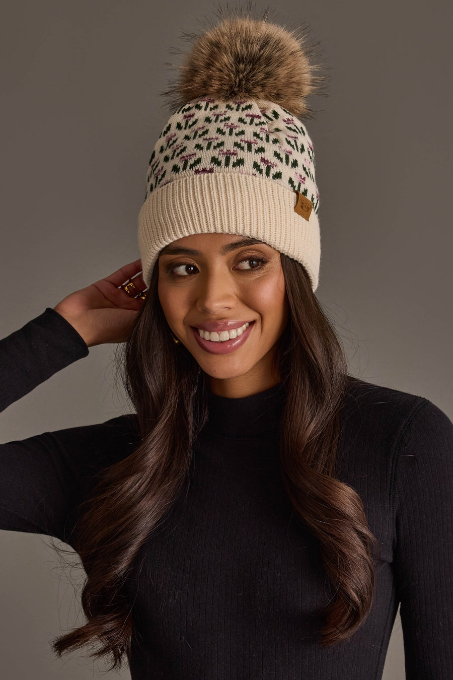 A woman in a black sweater wears the Panache Apparel Collection Cream Small Floral Pattern Knit Pom Hat, smiling and touching her hair against a neutral background.