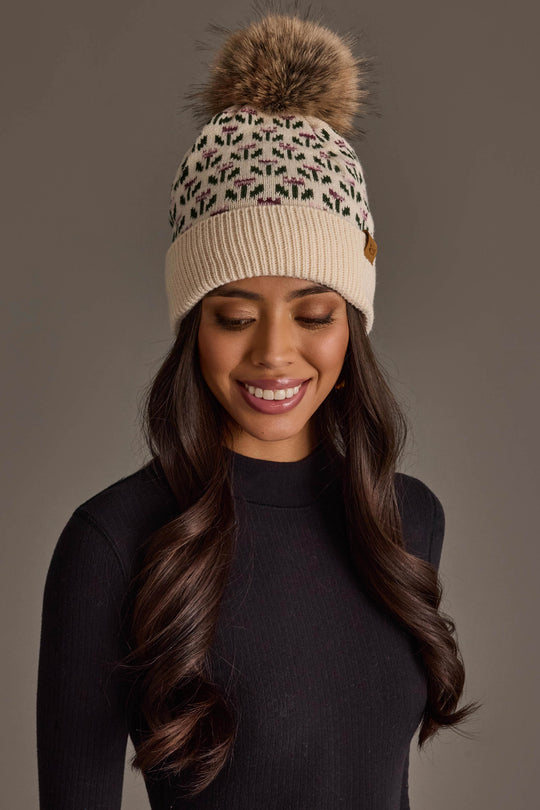 A woman with long dark hair smiles with her eyes closed against a plain background, wearing the Panache Apparel Collection Cream Small Floral Pattern Knit Pom Hat, a cream beanie featuring a green and pink floral pattern and faux fur pom-pom, paired with a black ribbed sweater.
