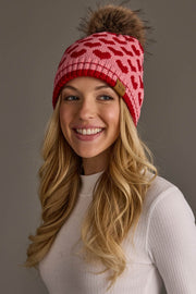 A woman with long blonde hair is smiling against a plain gray background, wearing a white ribbed top and the Panache Apparel Collection Pink & Red Heart Pattern Pom Hat, which features a red heart design and a brown pom-pom.