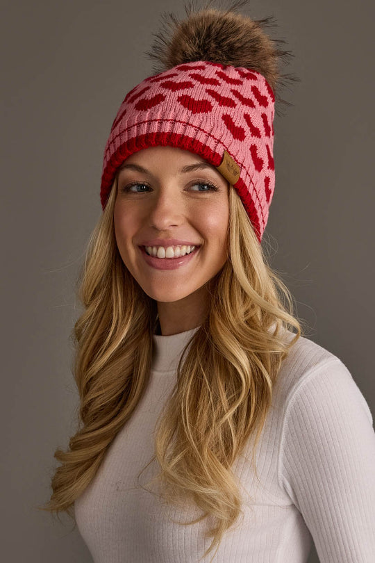 A woman with long blonde hair is smiling against a plain gray background, wearing a white ribbed top and the Panache Apparel Collection Pink & Red Heart Pattern Pom Hat, which features a red heart design and a brown pom-pom.