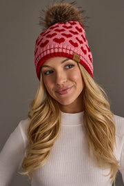 A woman with long blonde hair wears the Pink & Red Heart Pattern Pom Hat from Panache Apparel Collection, featuring a red heart design and a brown pom-pom. She is dressed in a white ribbed top and smiles softly against a neutral background.