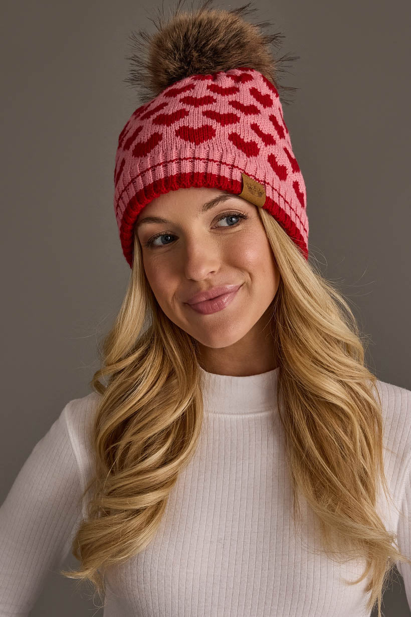 A woman with long blonde hair wears the Pink & Red Heart Pattern Pom Hat from Panache Apparel Collection, featuring a red heart design and a brown pom-pom. She is dressed in a white ribbed top and smiles softly against a neutral background.
