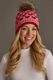 A smiling woman with long blonde hair, dressed in a white ribbed turtleneck, wears the Panache Apparel Collection Pink & Red Heart Pattern Pom Hat—featuring a playful red heart motif and a brown pom-pom—as she stands against a plain gray background.