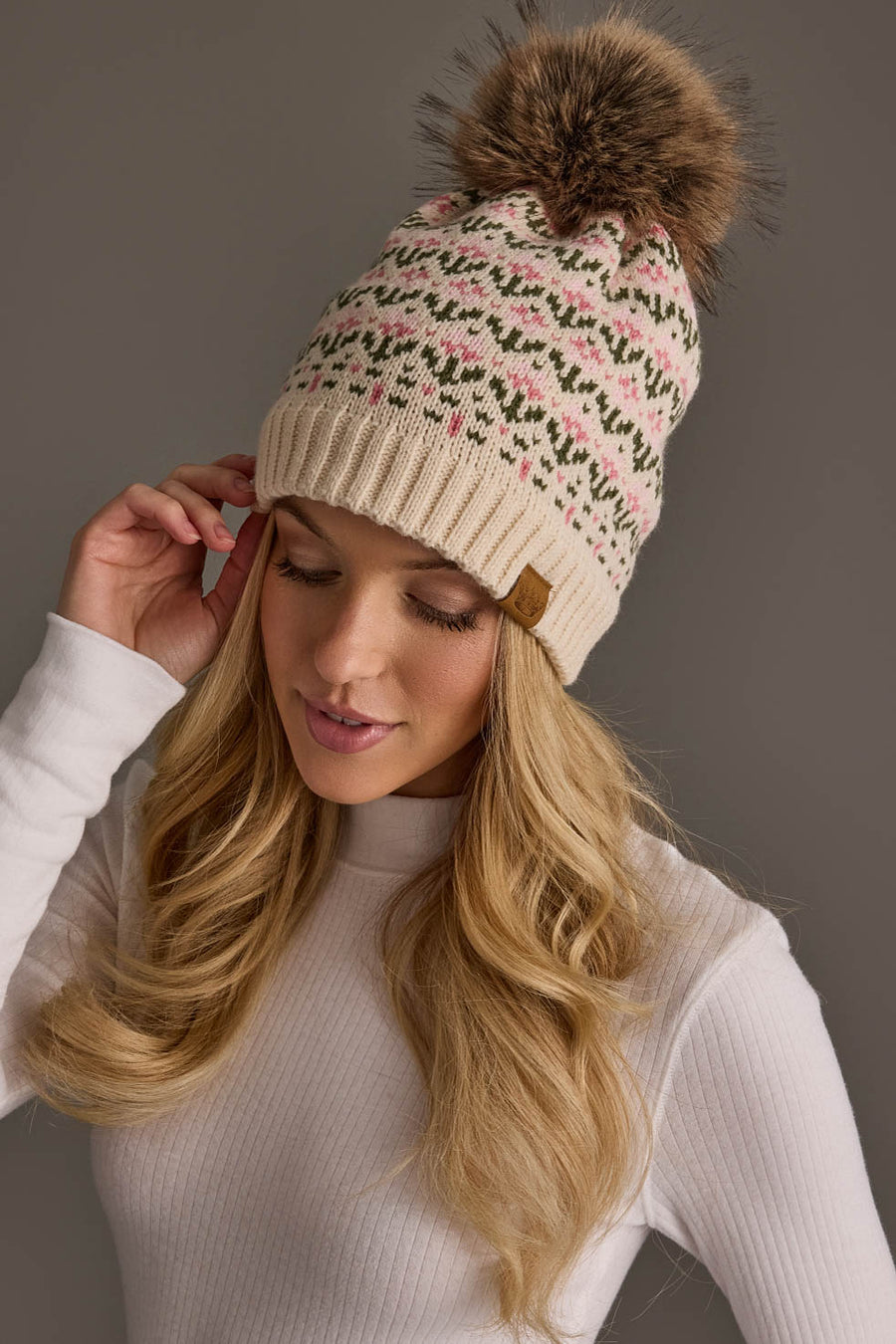 A woman with long blonde hair wears the Panache Apparel Collection Cream & Pink Floral Pattern Pom Hat, featuring a cream knit design with a floral pattern and a fur pom-pom. She is dressed in a white ribbed long-sleeve top and gently touches the side of her hat, looking down slightly.