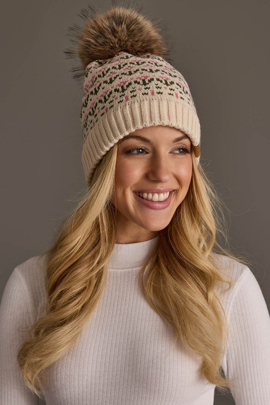 A smiling woman with long blonde hair wears the Panache Apparel Collection Cream & Pink Floral Pattern Pom Hat and a white ribbed long-sleeve top, posing against a plain gray background.